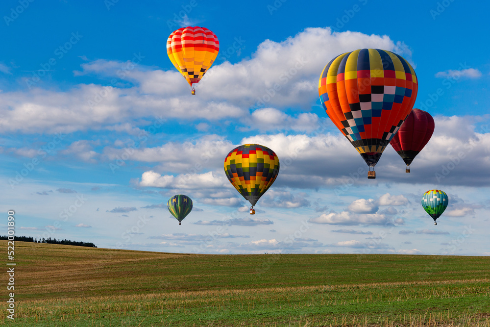 Obraz premium Multicolored hot air balloons fly in blue sky with white clouds over green field.