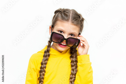 The child looks out from under sunglasses. A beautiful little girl in a yellow summer suit on a white isolated background.