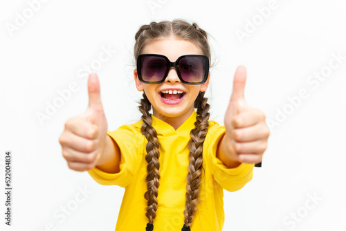 A teenager gives a thumbs up on a white isolated background. Portrait of a little girl in a yellow tracksuit and sunglasses.