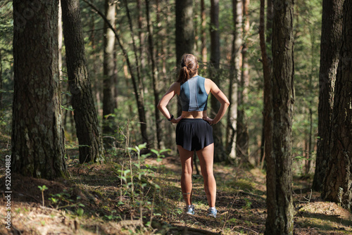 Rear view of female trail runner dressed in sportswear standing on trail in the forest and preparing to run.