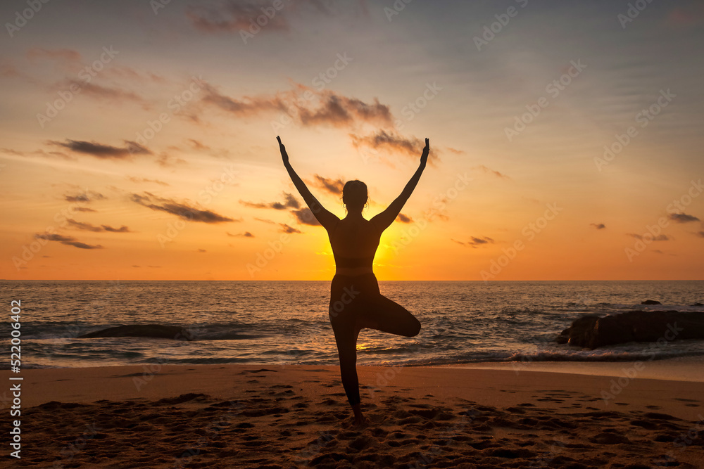 Slim woman does yoga position arms raised on tropical sea coast or ocean beach outdoors at sunset