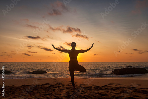 Silhouette slim woman does yoga position arms raised on tropical sea coast or ocean beach outdoors at sunset