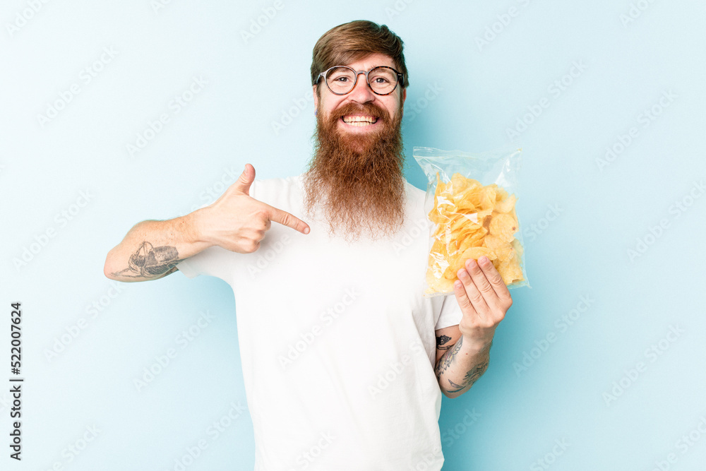 Young caucasian man holding a bag of chips isolated on blue background person pointing by hand to a shirt copy space, proud and confident Young caucasian man holding a bag of chips isolated on blue background person pointing by hand to a shirt copy space, proud and confident