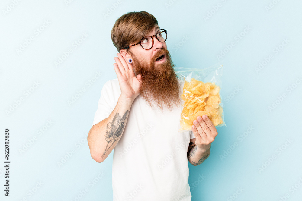 Young caucasian man holding a bag of chips isolated on blue background trying to listening a gossip. Young caucasian man holding a bag of chips isolated on blue background trying to listening a gossip.