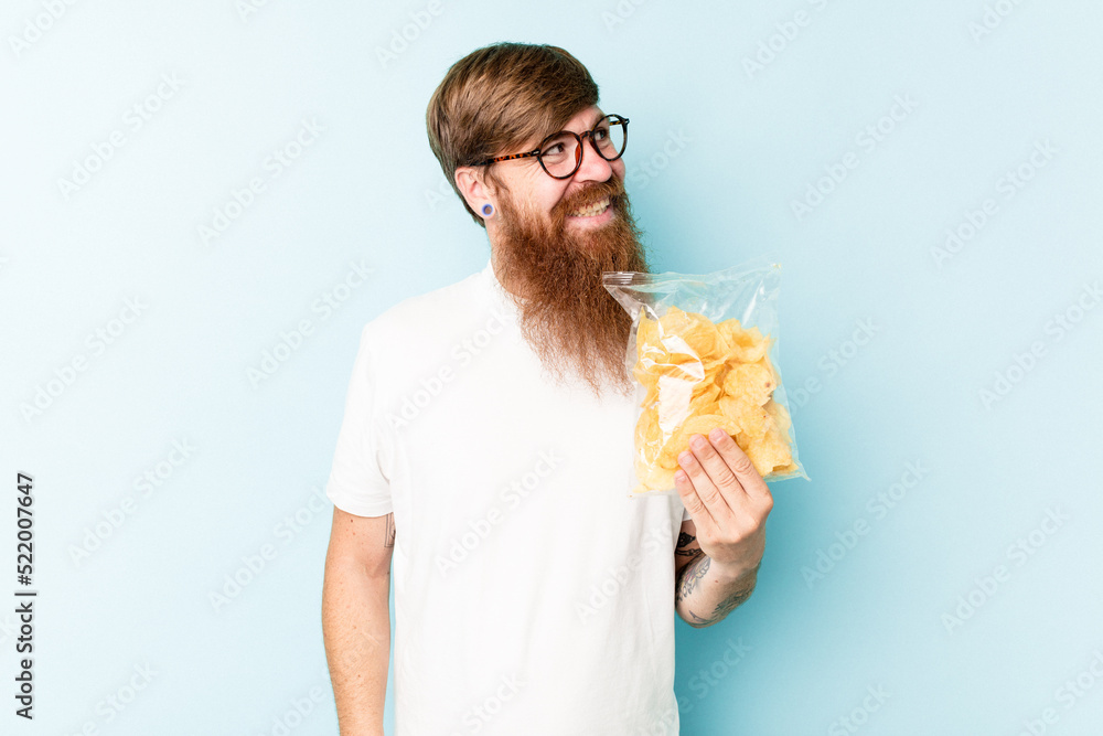 Young caucasian man holding a bag of chips isolated on blue background looks aside smiling, cheerful and pleasant. Young caucasian man holding a bag of chips isolated on blue background looks aside smiling, cheerful and pleasant.