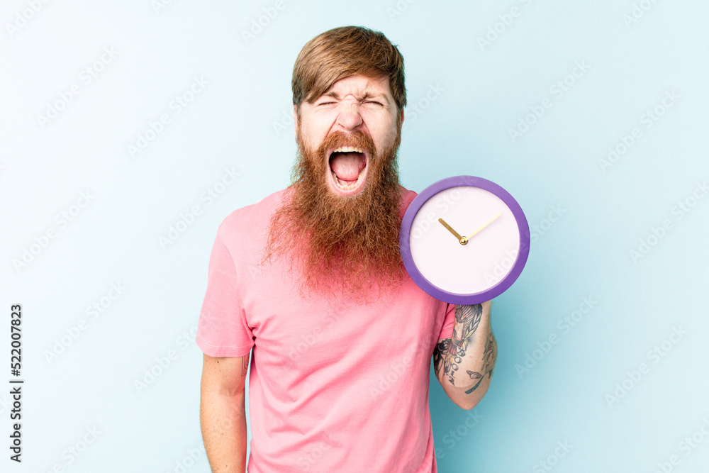 Young caucasian man holding a clock isolated on blue background screaming very angry and aggressive. Young caucasian man holding a clock isolated on blue background screaming very angry and aggressive.