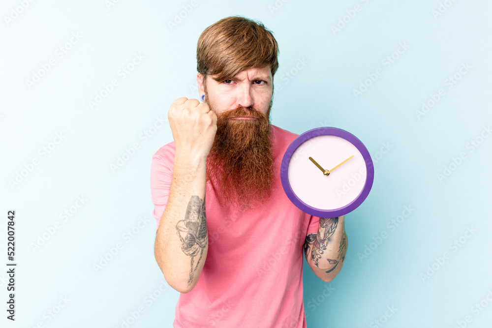 Young caucasian man holding a clock isolated on blue background showing fist to camera, aggressive facial expression. Young caucasian man holding a clock isolated on blue background showing fist to camera, aggressive facial expression.