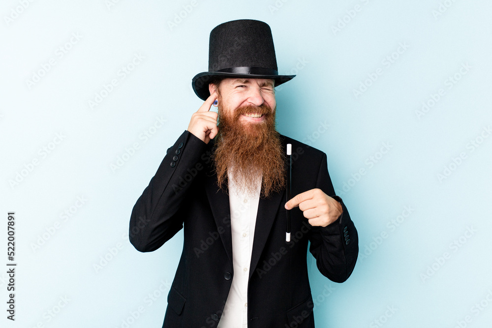 Young magician man isolated on blue background covering ears with hands. Young magician man isolated on blue background covering ears with hands.