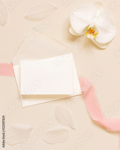 Wedding card over envelope near white orchid flower and silk ribbons on yellow, mockup