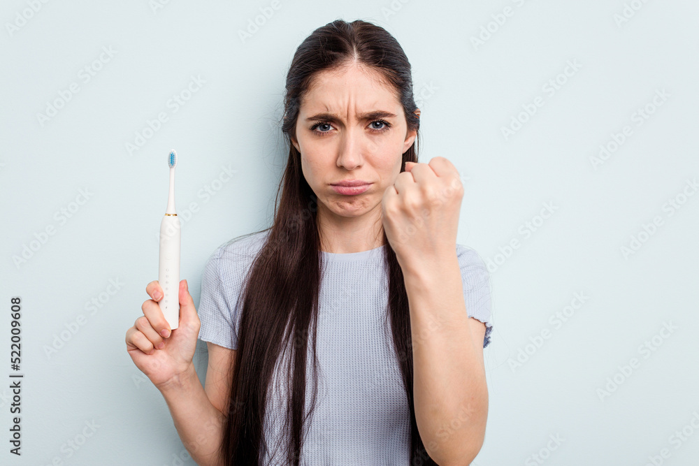 Young caucasian woman holding a electric toothbrush isolated on blue background showing fist to camera, aggressive facial expression. Young caucasian woman holding a electric toothbrush isolated on blue background showing fist to camera, aggressive facial expression.