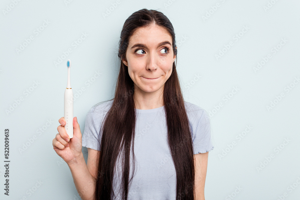 Young caucasian woman holding a electric toothbrush isolated on blue background confused, feels doubtful and unsure. Young caucasian woman holding a electric toothbrush isolated on blue background confused, feels doubtful and unsure.