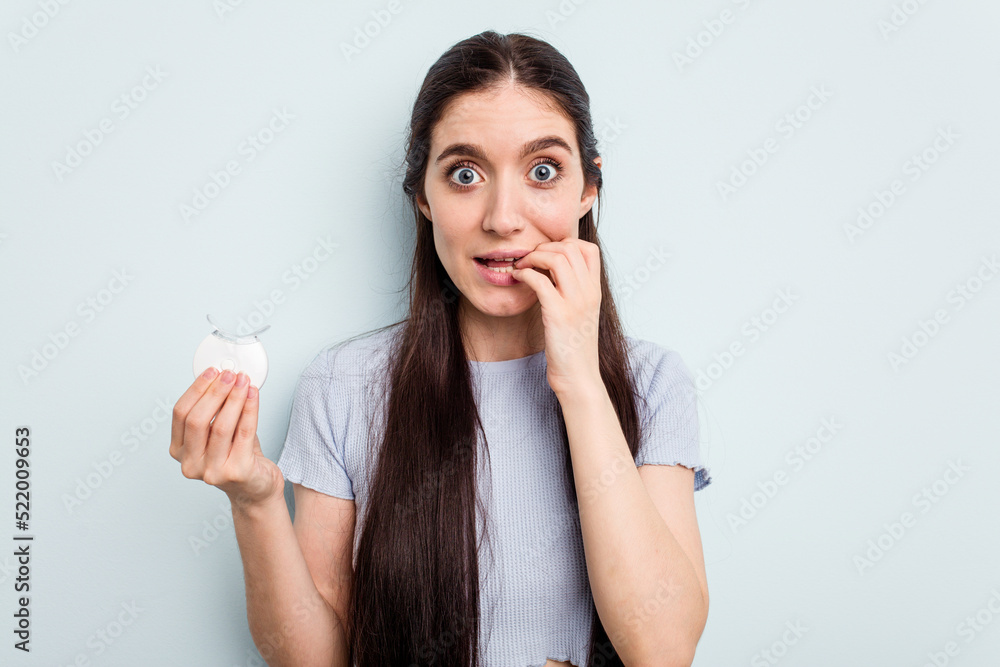 Young caucasian woman holding teeth whitener isolated on blue background biting fingernails, nervous and very anxious. Young caucasian woman holding teeth whitener isolated on blue background biting fingernails, nervous and very anxious.