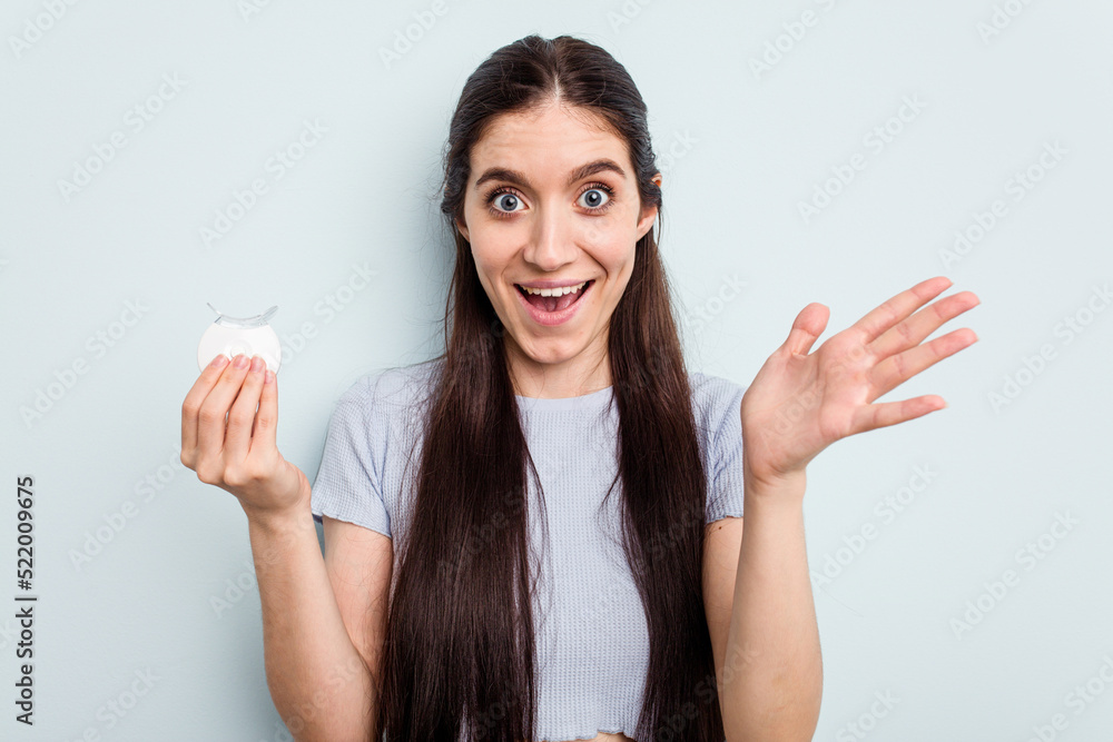Young caucasian woman holding teeth whitener isolated on blue background receiving a pleasant surprise, excited and raising hands.