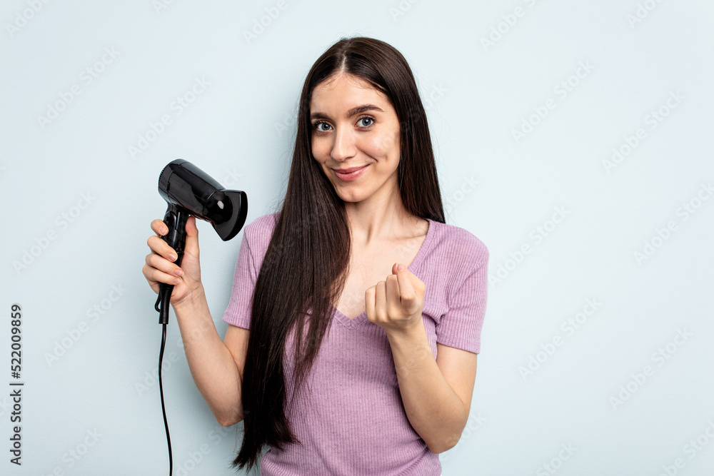 Young caucasian woman holding hairdryer isolated on blue background pointing with finger at you as if inviting come closer. Young caucasian woman holding hairdryer isolated on blue background pointing with finger at you as if inviting come closer.