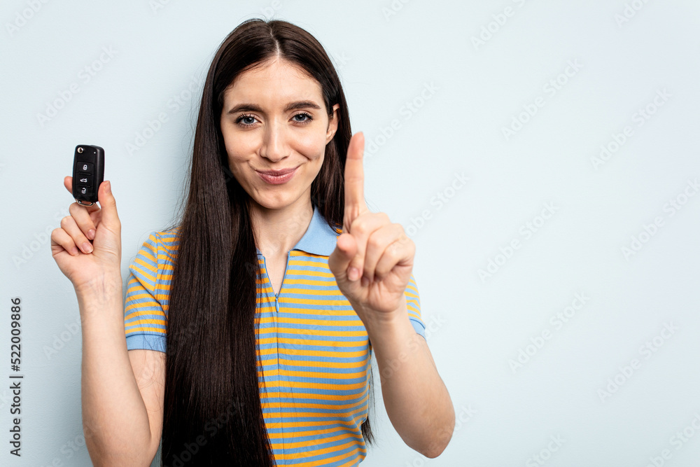 Young caucasian woman holding car keys isolated on blue background showing number one with finger. Young caucasian woman holding car keys isolated on blue background showing number one with finger.