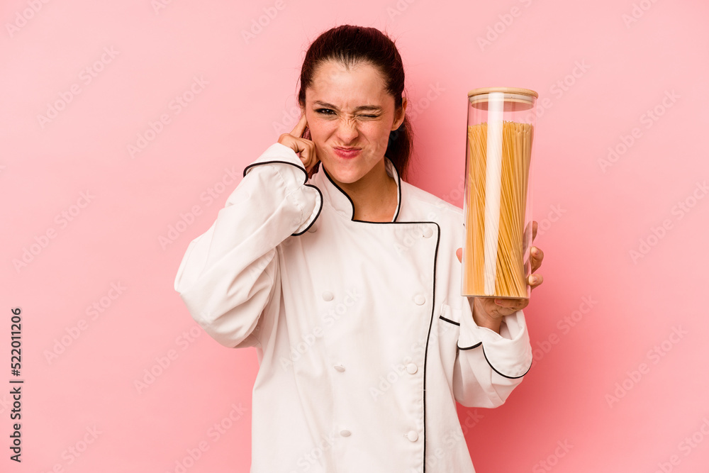 Young chef caucasian woman holding a spaghettis jar isolated on pink background covering ears with hands. Young chef caucasian woman holding a spaghettis jar isolated on pink background covering ears with hands.