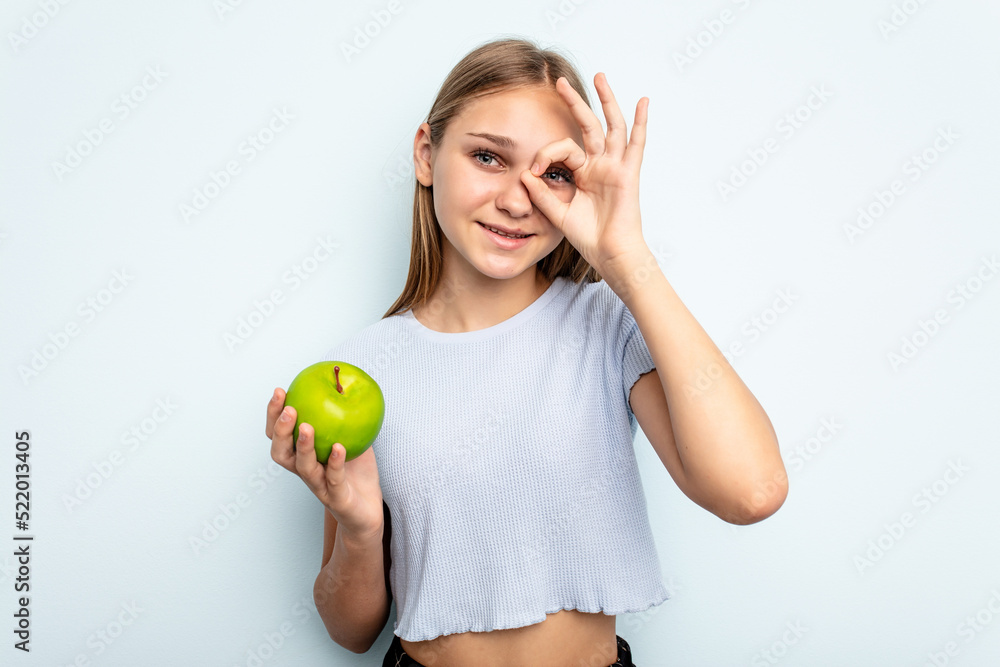 Young caucasian girl holding an apple isolated on blue background excited keeping ok gesture on eye. Young caucasian girl holding an apple isolated on blue background excited keeping ok gesture on eye.