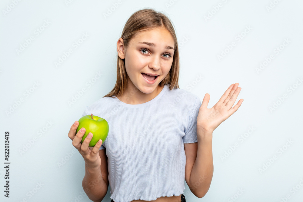 Young caucasian girl holding an apple isolated on blue background receiving a pleasant surprise, excited and raising hands. Young caucasian girl holding an apple isolated on blue background receiving a pleasant surprise, excited and raising hands.