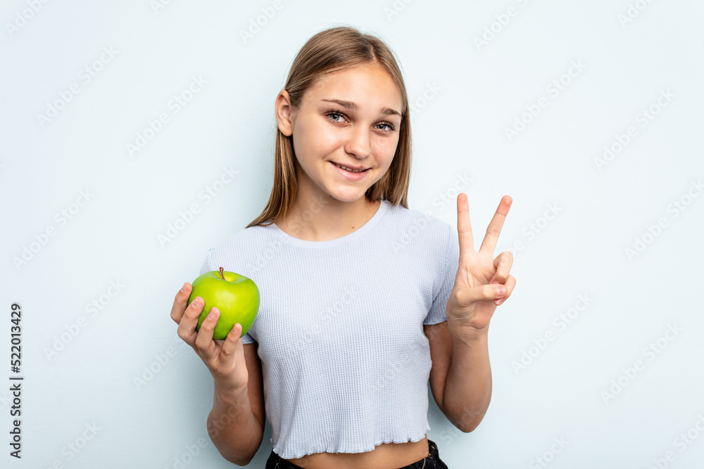 Young caucasian girl holding an apple isolated on blue background showing number two with fingers. Young caucasian girl holding an apple isolated on blue background showing number two with fingers.