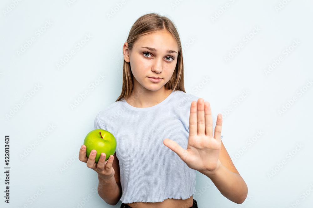 Young caucasian girl holding an apple isolated on blue background standing with outstretched hand showing stop sign, preventing you. Young caucasian girl holding an apple isolated on blue background standing with outstretched hand showing stop sign, preventing you.