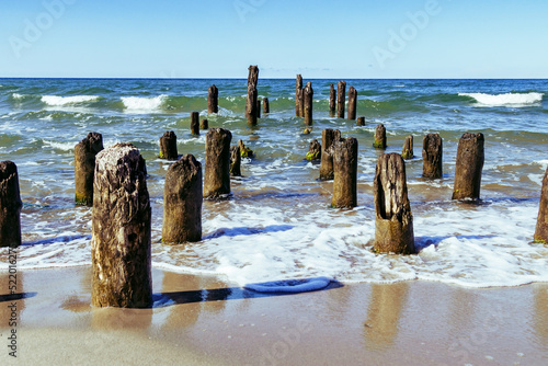 Seascape, row of wooden piles on sandy beach, blue sky, blue sea water of Baltic sea, Filinskaya bay beach, Russia. .Old weathered wooden poles in water. Summer day, waves and wind