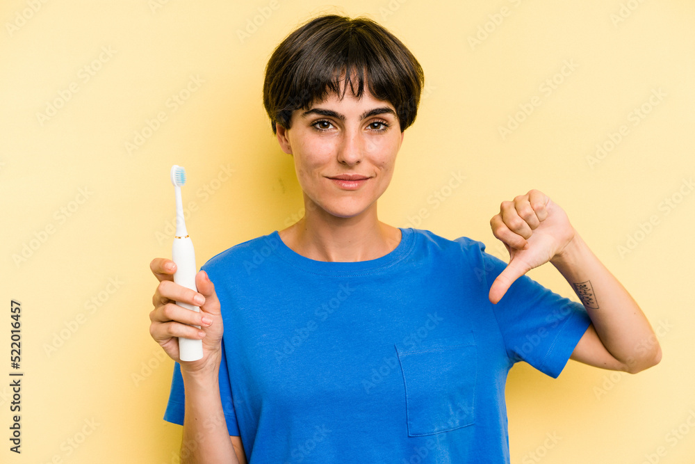 Young caucasian woman holding a electric toothbrush isolated on yellow background showing a dislike gesture, thumbs down. Disagreement concept. Young caucasian woman holding a electric toothbrush isolated on yellow background showing a dislike gesture, thumbs down. Disagreement concept.