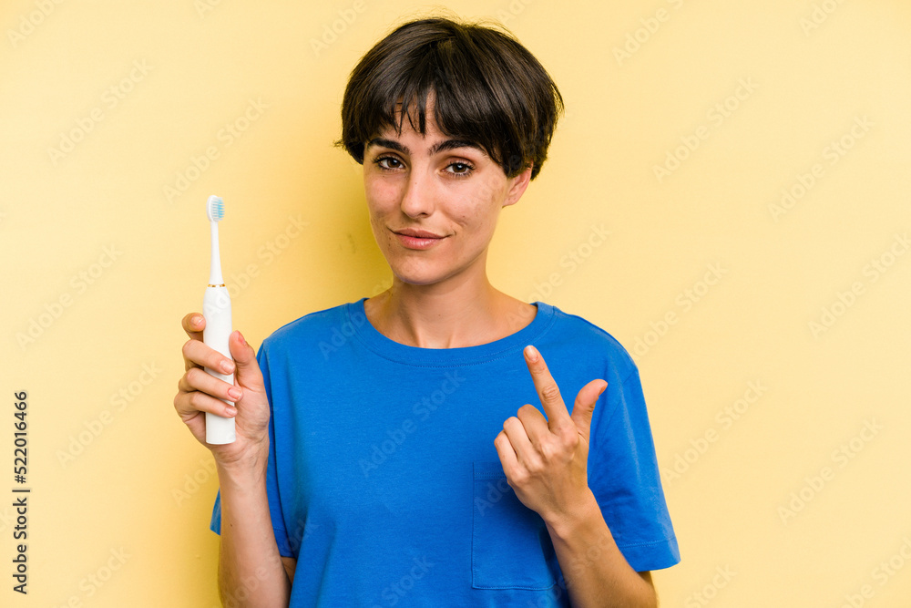 Young caucasian woman holding a electric toothbrush isolated on yellow background pointing with finger at you as if inviting come closer. Young caucasian woman holding a electric toothbrush isolated on yellow background pointing with finger at you as if inviting come closer.