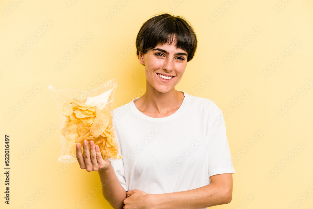 Young caucasian man holding crisps isolated on yellow background laughing and having fun. Young caucasian man holding crisps isolated on yellow background laughing and having fun.