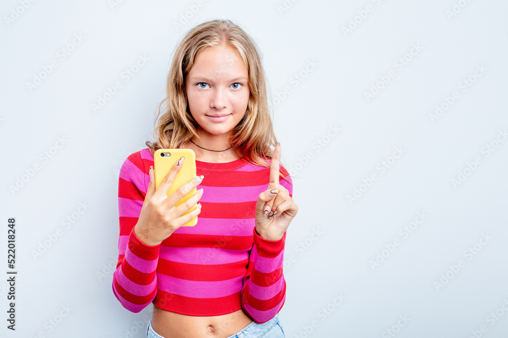 Caucasian teen girl holding mobile phone isolated on blue background showing number one with finger. Caucasian teen girl holding mobile phone isolated on blue background showing number one with finger.