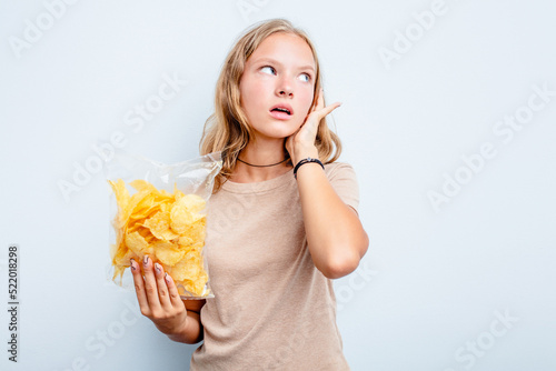 Caucasian teen girl holding bag of chips isolated on blue background trying to listening a gossip.