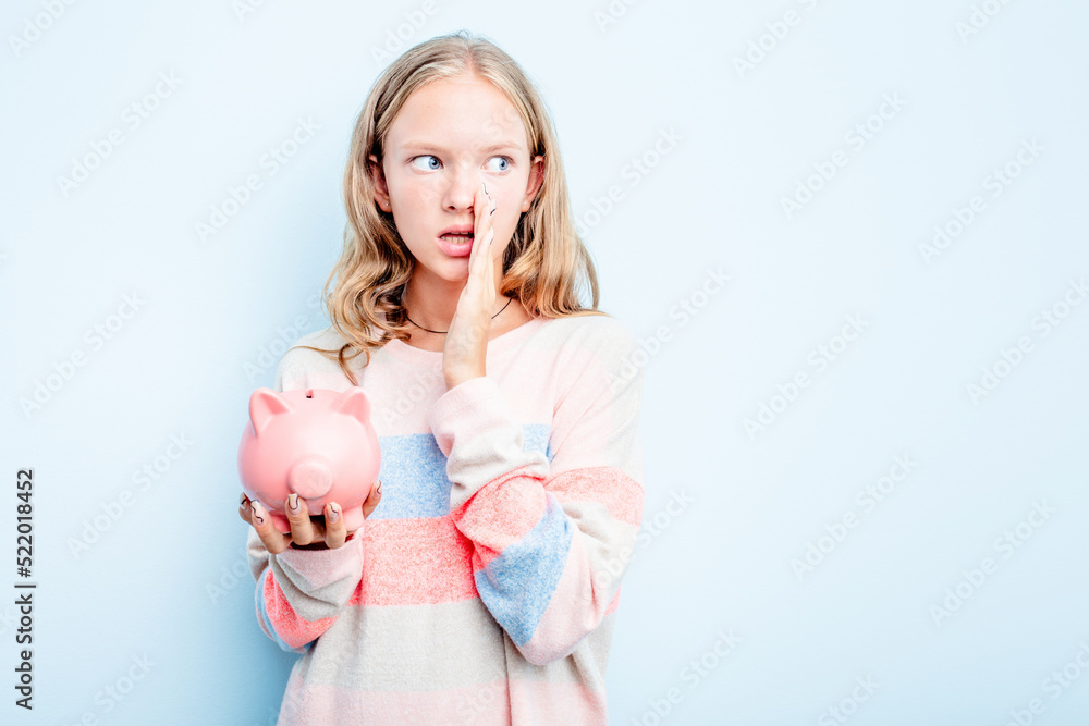 Caucasian teen girl holding a piggybank isolated on blue background is saying a secret hot braking news and looking aside Caucasian teen girl holding a piggybank isolated on blue background is saying a secret hot braking news and looking aside