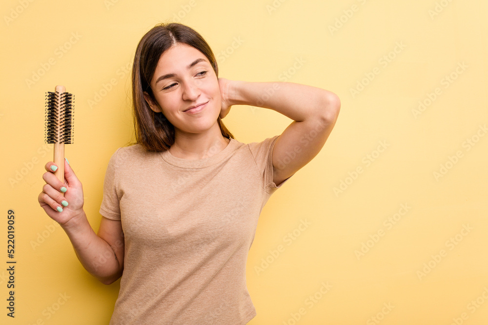 Young caucasian woman holding a brush hair isolated on yellow background touching back of head, thinking and making a choice. Young caucasian woman holding a brush hair isolated on yellow background touching back of head, thinking and making a choice.