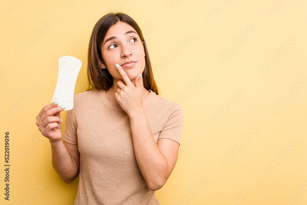 Young caucasian woman holding a sanitary napkin isolated on yellow background looking sideways with doubtful and skeptical expression. Young caucasian woman holding a sanitary napkin isolated on yellow background looking sideways with doubtful and skeptical expression.
