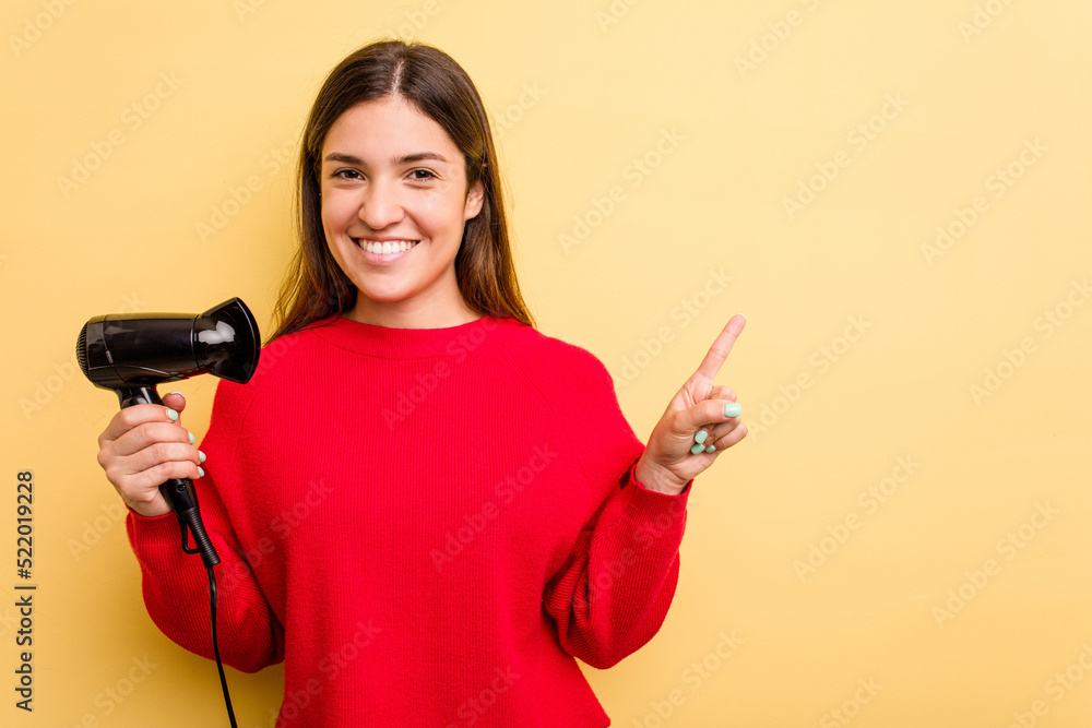 Young caucasian woman holding hairdryer isolated on yellow background smiling and pointing aside, showing something at blank space. Young caucasian woman holding hairdryer isolated on yellow background smiling and pointing aside, showing something at blank space.
