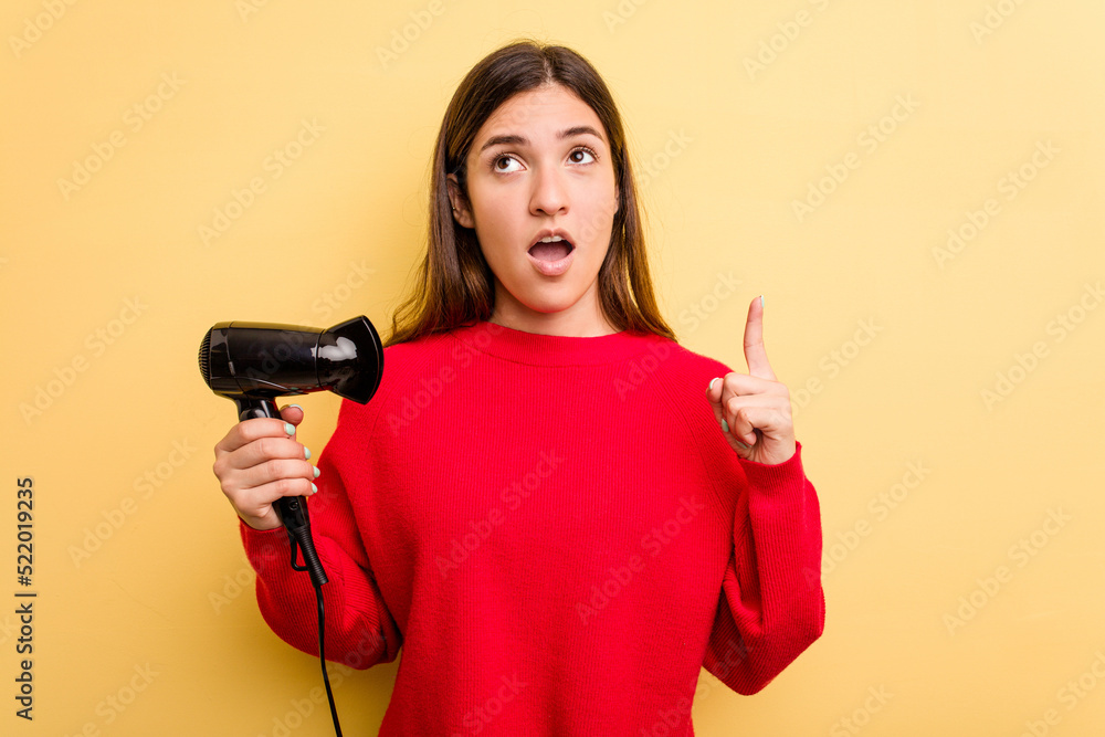 Young caucasian woman holding hairdryer isolated on yellow background pointing upside with opened mouth. Young caucasian woman holding hairdryer isolated on yellow background pointing upside with opened mouth.