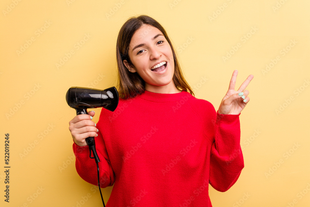 Young caucasian woman holding hairdryer isolated on yellow background joyful and carefree showing a peace symbol with fingers. Young caucasian woman holding hairdryer isolated on yellow background joyful and carefree showing a peace symbol with fingers.