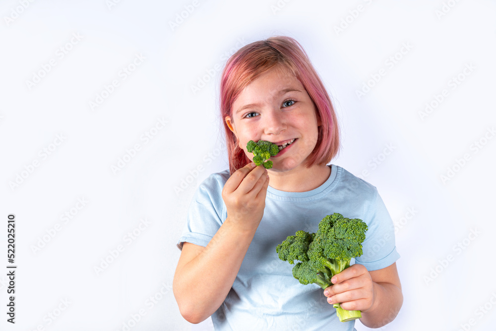 Concept of healthy baby children food, eat vegetables. Cute attractive european blonde kid girl eating fresh green broccoli, on gray background Concept of healthy baby children food, eat vegetables. Cute attractive european blonde kid girl eating fresh green broccoli, on gray background
