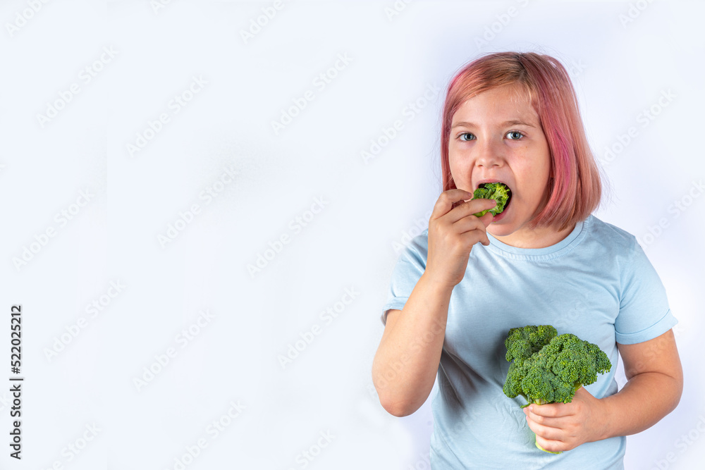 Concept of healthy baby children food, eat vegetables. Cute attractive european blonde kid  girl eating fresh green broccoli, on gray background 