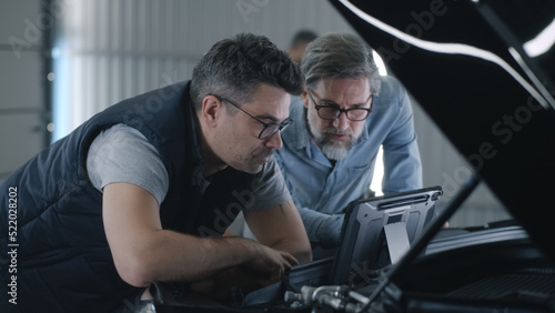 A man mechanic in glasses and a client standing together near the opened hood in a car service, and discussing the results of car diagnostics using a digital tablet computer