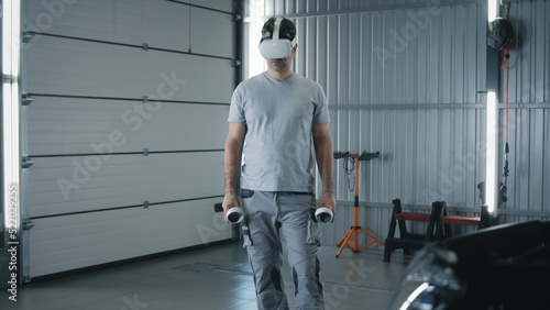 Man worker in uniform in a modern virtual reality glasses with controllers checking and testing a car after repairing in a car service