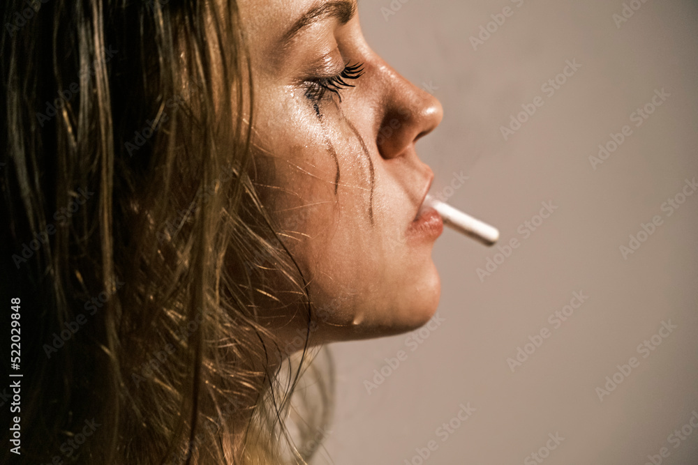 Fototapeta premium Crying young woman with cigarette in her mouth. Portrait of side view of unhappy woman experiencing an emotional crisis in her life. Domestic psychological and physical violence