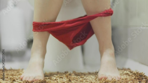 Woman is sitting on the toilet in a home bathroom, taking off her red panties and scratching her leg. Close-up of feet. The concept of constipation and diarrhea. photo