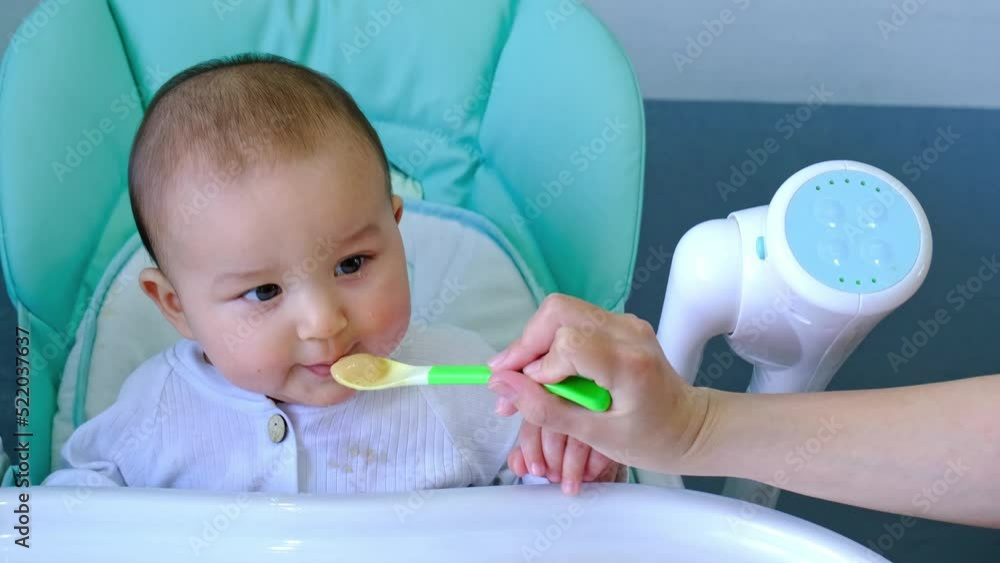Mom feeds the baby with a spoon of vegetable puree at the children's feeding table. Baby's appetite, healthy nutrition, introduction of complementary foods. Copyspace, mock up Mom feeds the baby with a spoon of vegetable puree at the children's feeding table. Baby's appetite, healthy nutrition, introduction of complementary foods. Copyspace, mock up