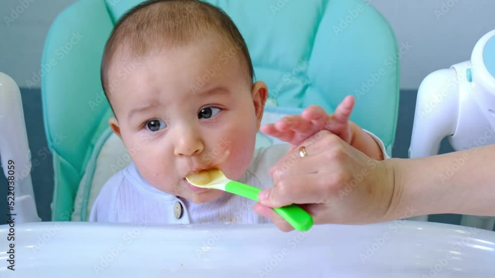 Mom feeds the baby with a spoon of vegetable puree at the children's feeding table. Baby's appetite, healthy nutrition, introduction of complementary foods. Copyspace, mock up Mom feeds the baby with a spoon of vegetable puree at the children's feeding table. Baby's appetite, healthy nutrition, introduction of complementary foods. Copyspace, mock up