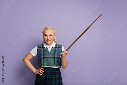Smiling blonde woman in vest holding pointer and hand on hip on purple background
