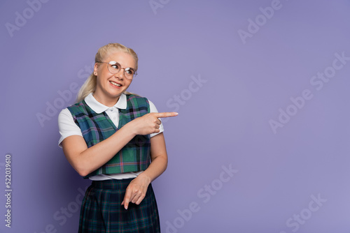 Smiling student in uniform and eyeglasses pointing with finger isolated on purple