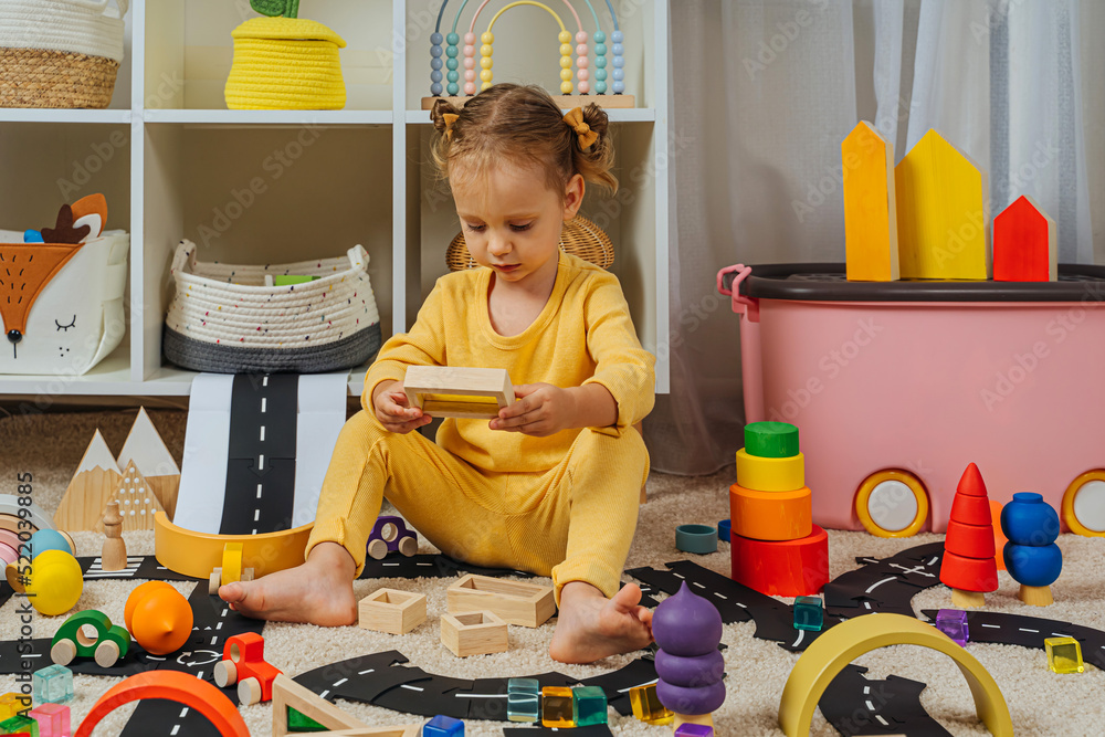 A little girl sitting on floor plays with toy wooden blocks cars and