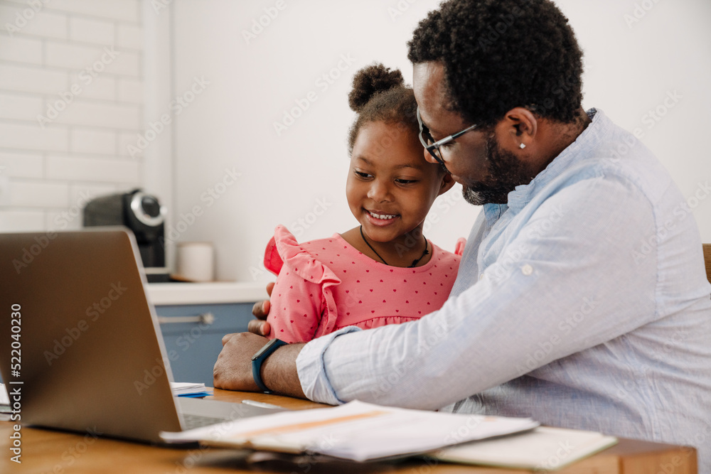 African american man hugging daughter while working on laptop at home African american man hugging daughter while working on laptop at home