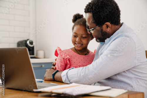 African american man hugging daughter while working on laptop at home