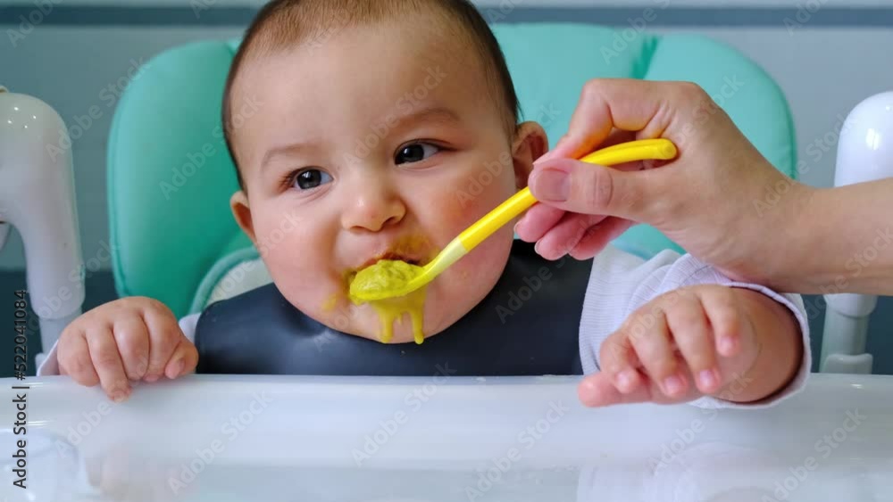 Mom feeds the baby with a spoon of vegetable puree at the children's feeding table. Baby's appetite, healthy nutrition, introduction of complementary foods. Copyspace, mock up Mom feeds the baby with a spoon of vegetable puree at the children's feeding table. Baby's appetite, healthy nutrition, introduction of complementary foods. Copyspace, mock up